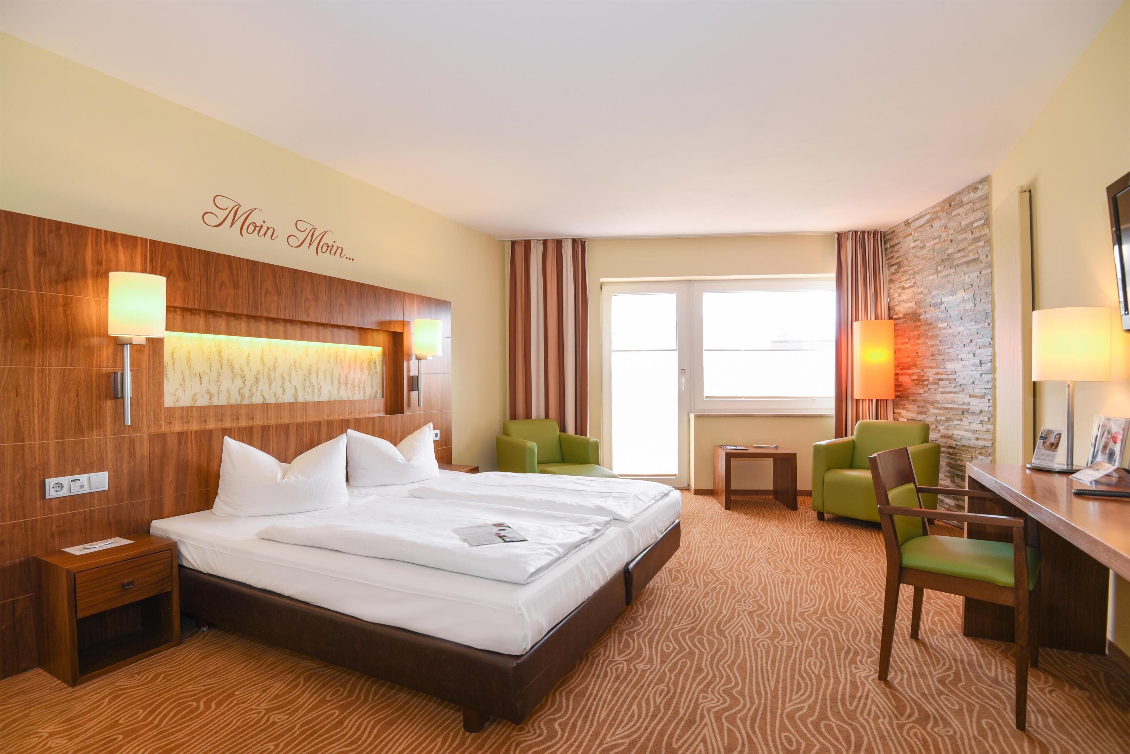 A hotel room with a large bed, two green chairs, a desk, and a stone accent wall.