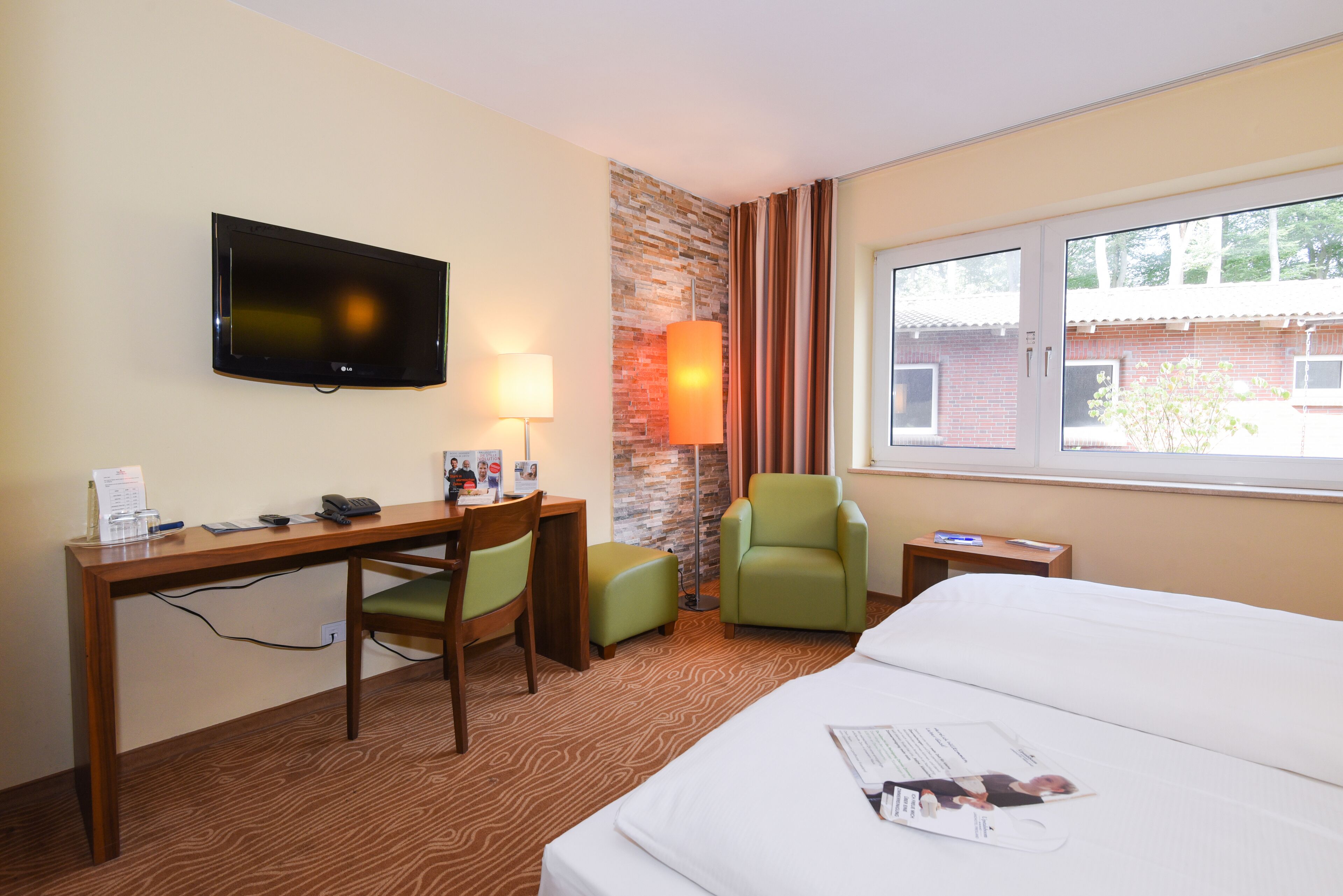 Ein Hotelzimmer mit Bett, Schreibtisch, Stuhl, Fernseher, Lampe und einem Fenster mit Blick auf ein Gebäude.