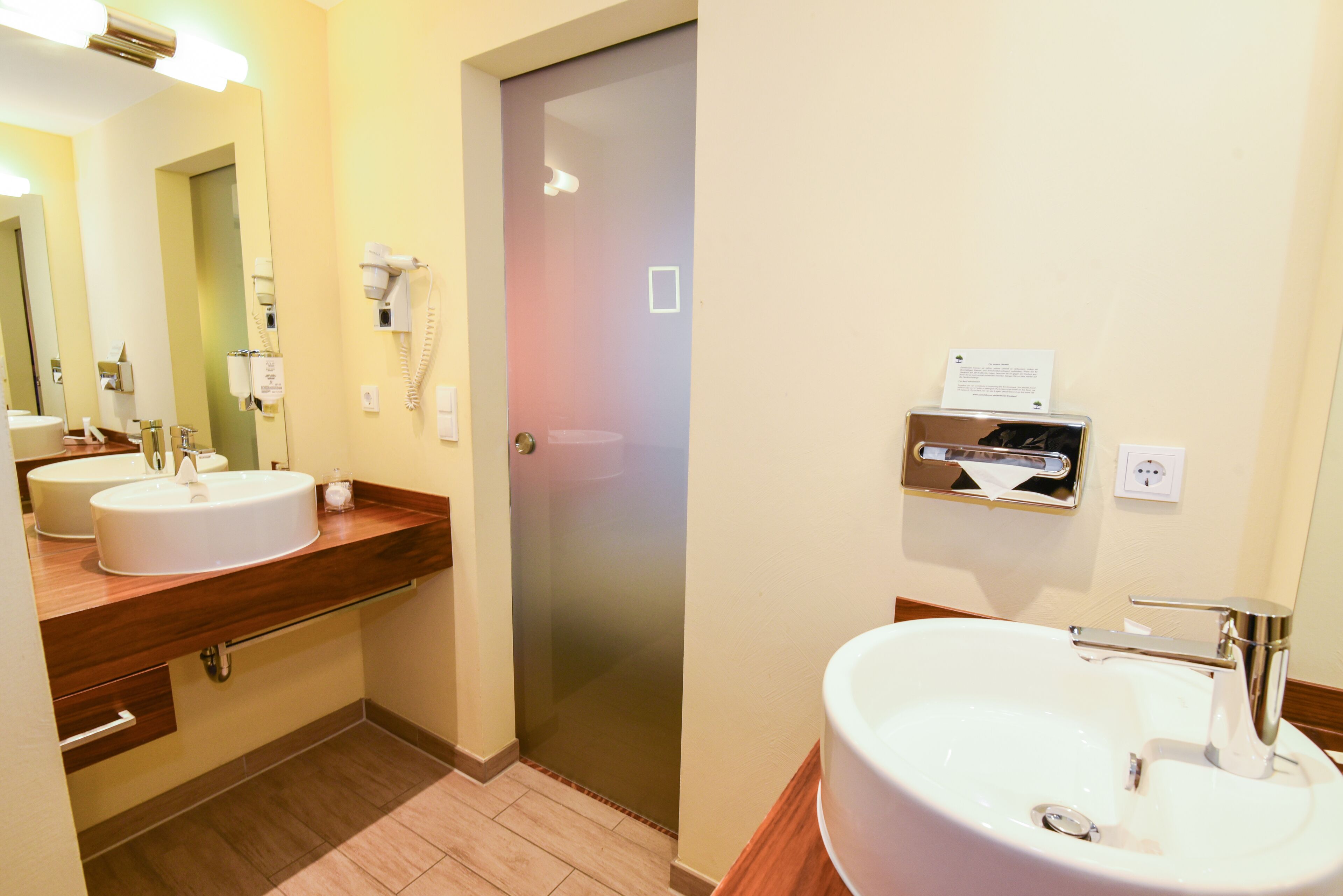 A hotel bathroom with two sinks, a hairdryer, and a towel dispenser.