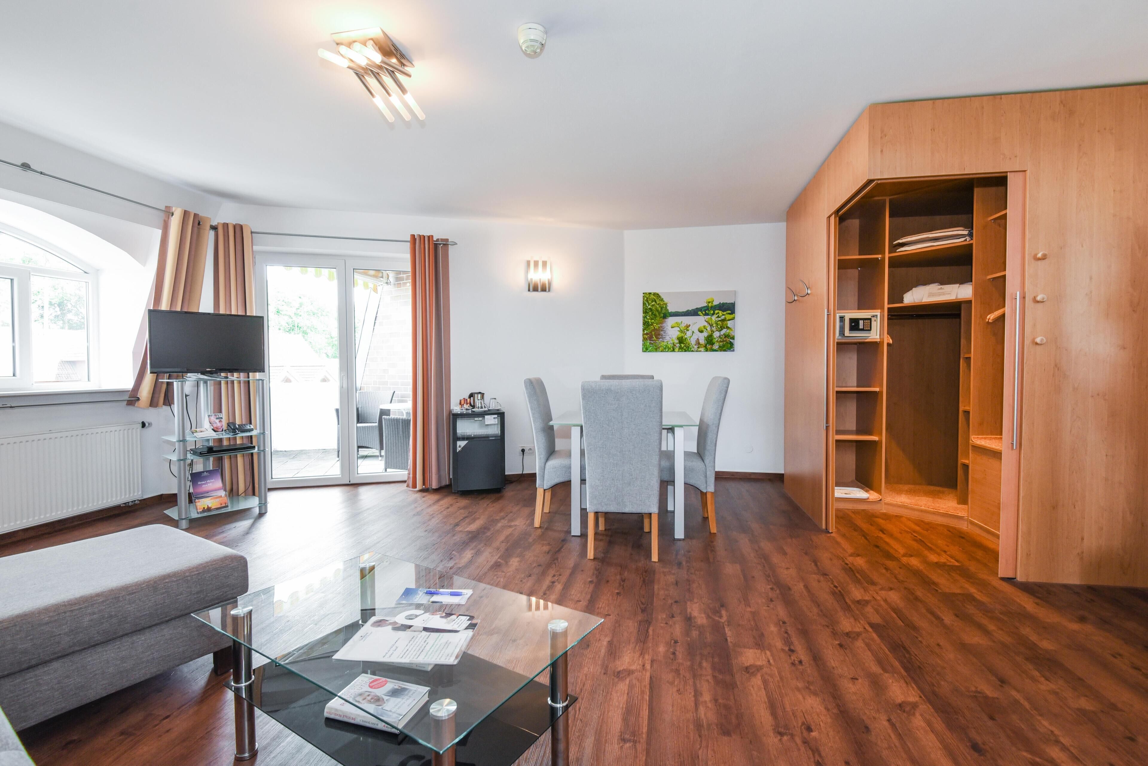 Una sala de estar moderna con una mesa de centro de vidrio, un sofá, un televisor, un comedor con sillas, un armario de madera y un ventanal grande.