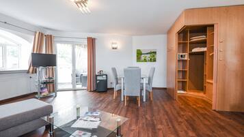 Una sala de estar moderna con una mesa de centro de vidrio, un sofá, un televisor, un comedor con sillas, un armario de madera y un ventanal grande.