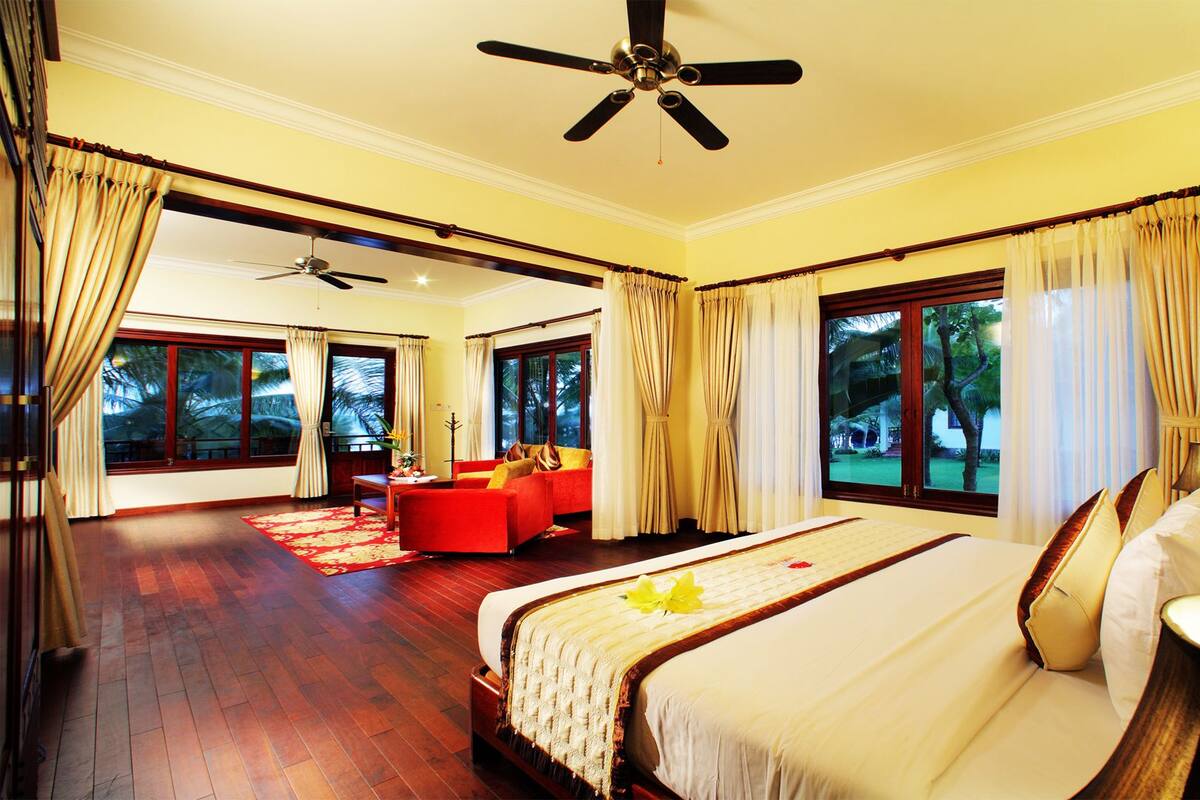 A spacious bedroom with a large bed, a sitting area with a red sofa, and a view of the outdoors through large windows.