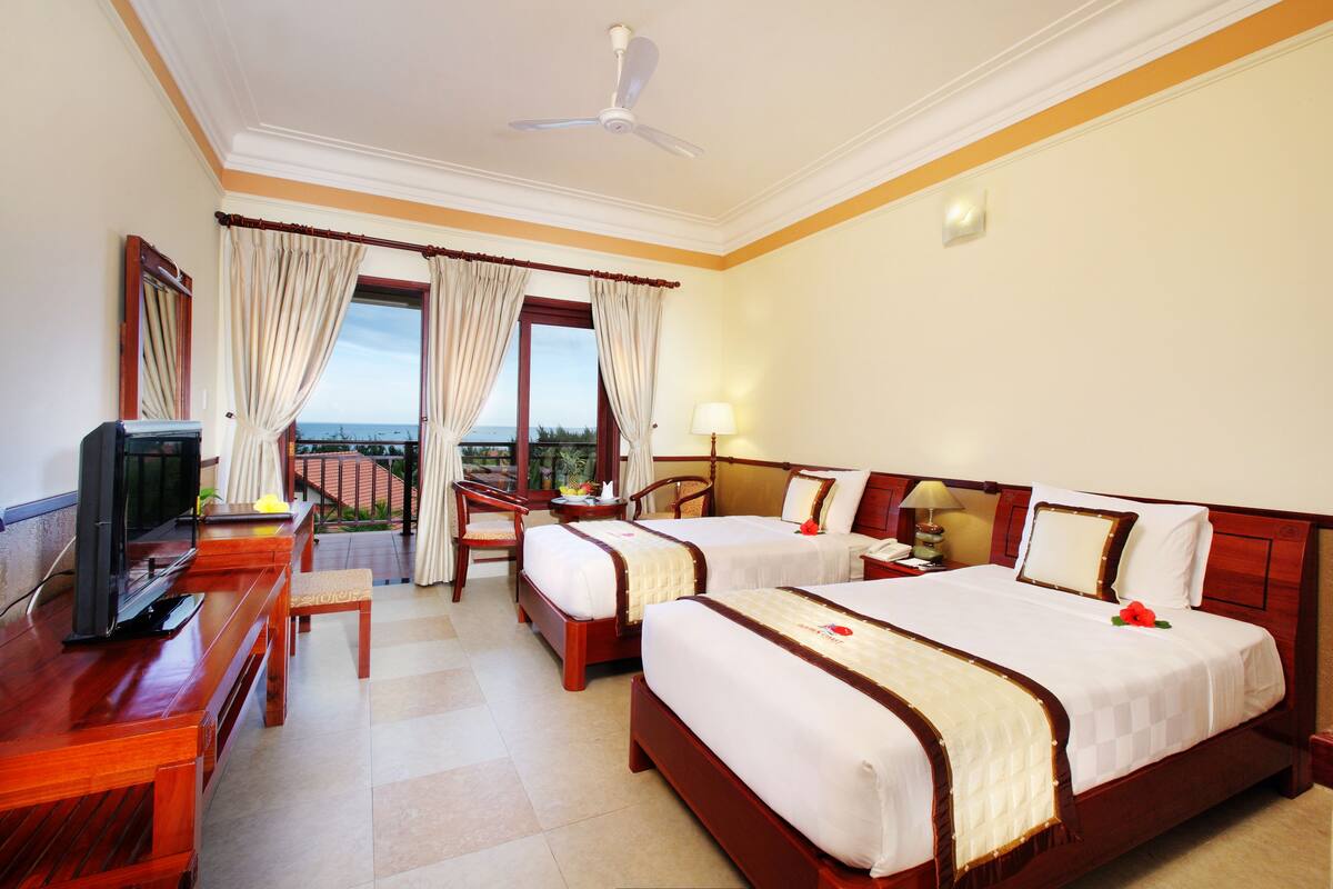 A hotel room with two beds, a desk with a television, a chair, and a view of the sea.