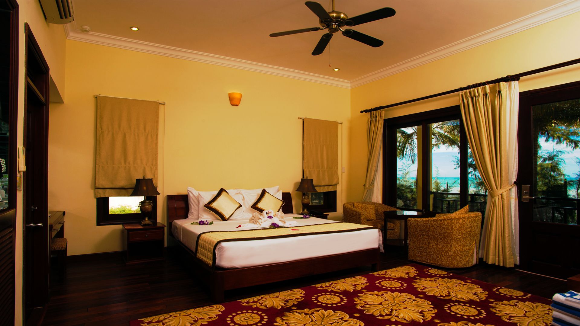 A hotel room with a bed, a ceiling fan, a window with curtains, a view of the beach, and a patterned carpet.