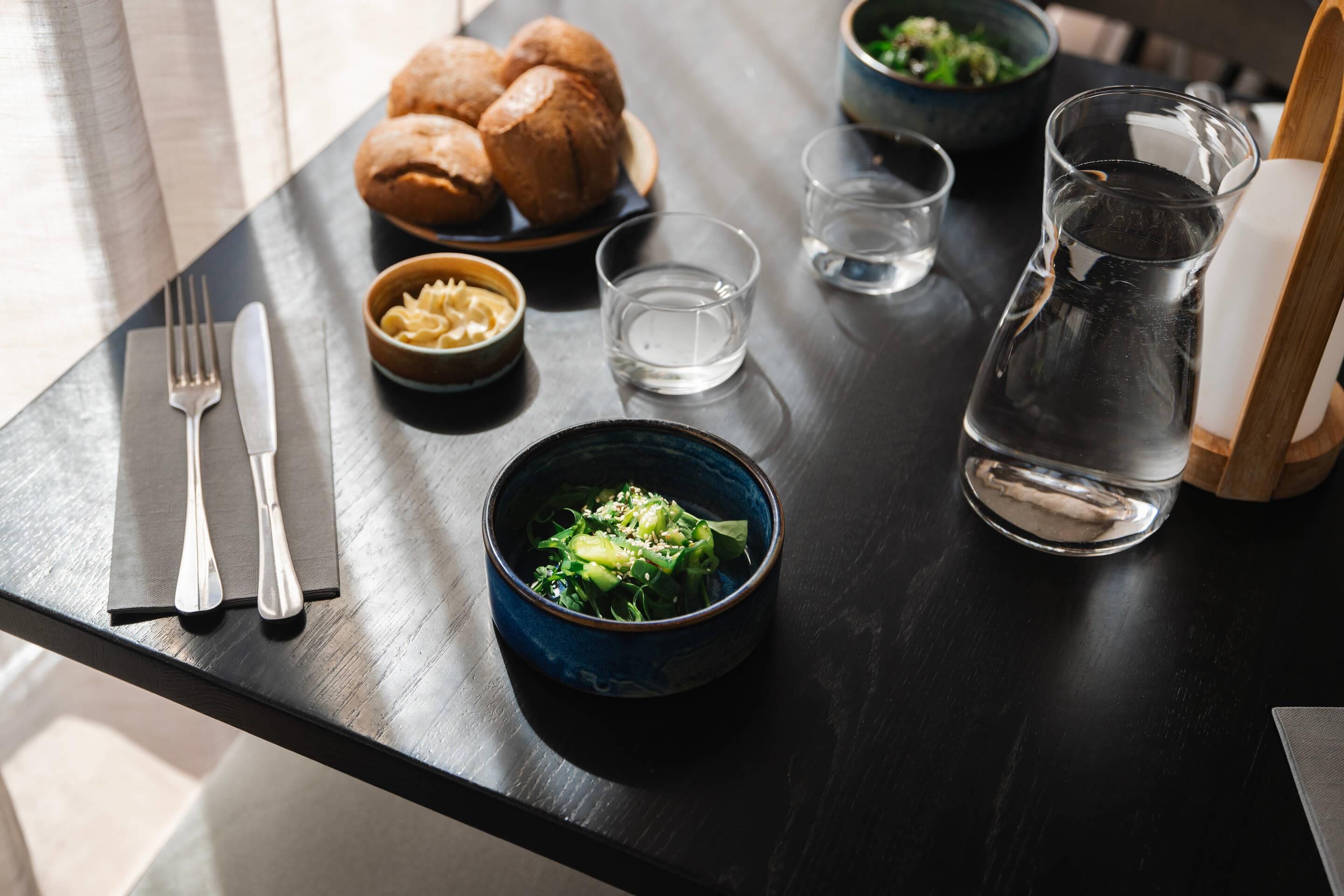 A dining table with a dark wooden surface, a bowl of pasta, a bowl of salad, a pitcher of water, a plate of bread, and a set of cutlery.