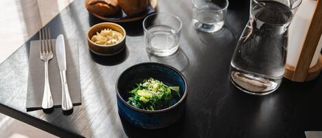 A dining table with a dark wooden surface, a bowl of pasta, a bowl of salad, a pitcher of water, a plate of bread, and a set of cutlery.