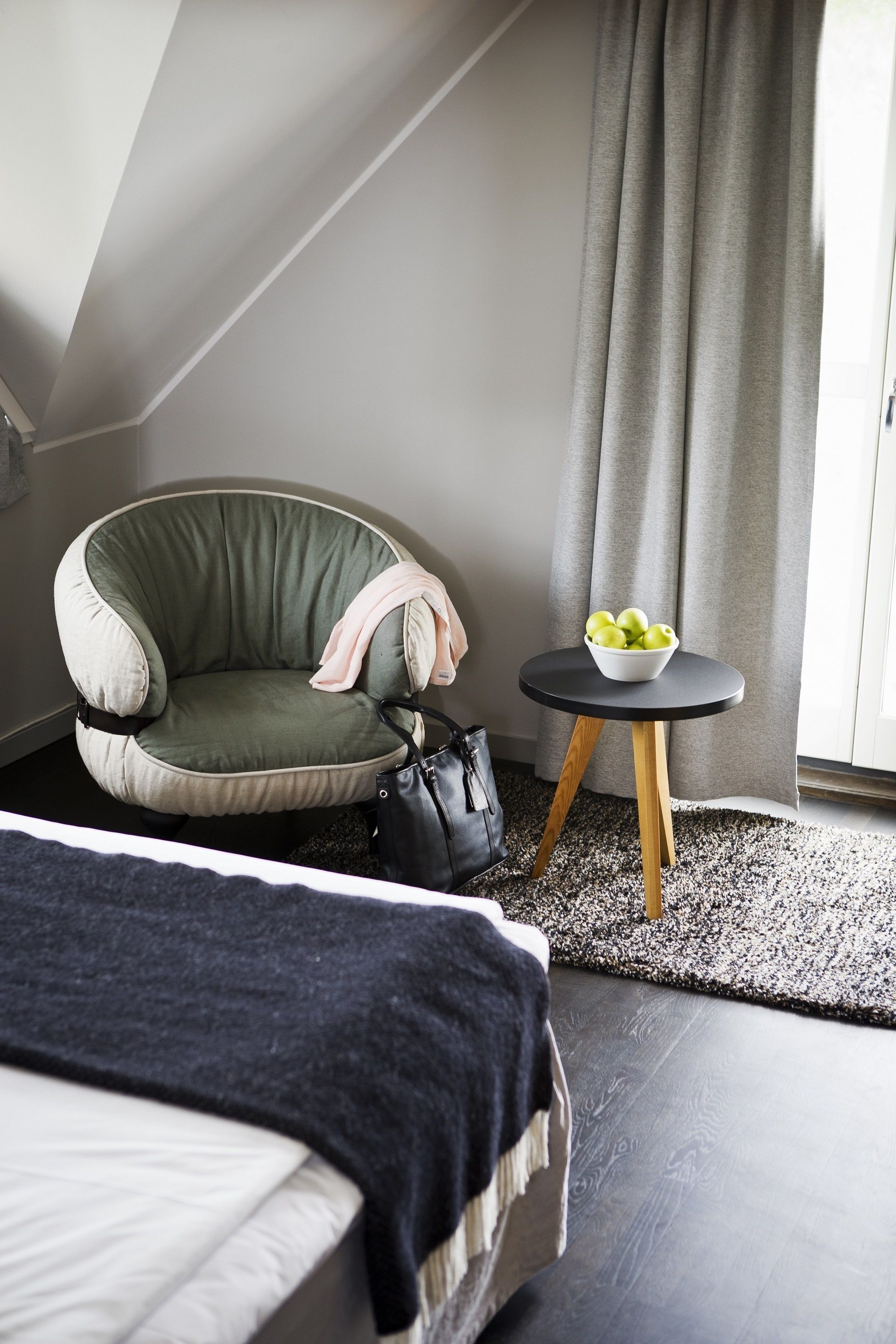 A modern hotel room with a curved green armchair, a small round table with a bowl of fruit, a black handbag, and a bed with a dark blanket.