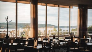 A restaurant with large windows offering a view of a lake and hills. Tables and chairs are arranged indoors. - Selma Spa+ (Sunne)