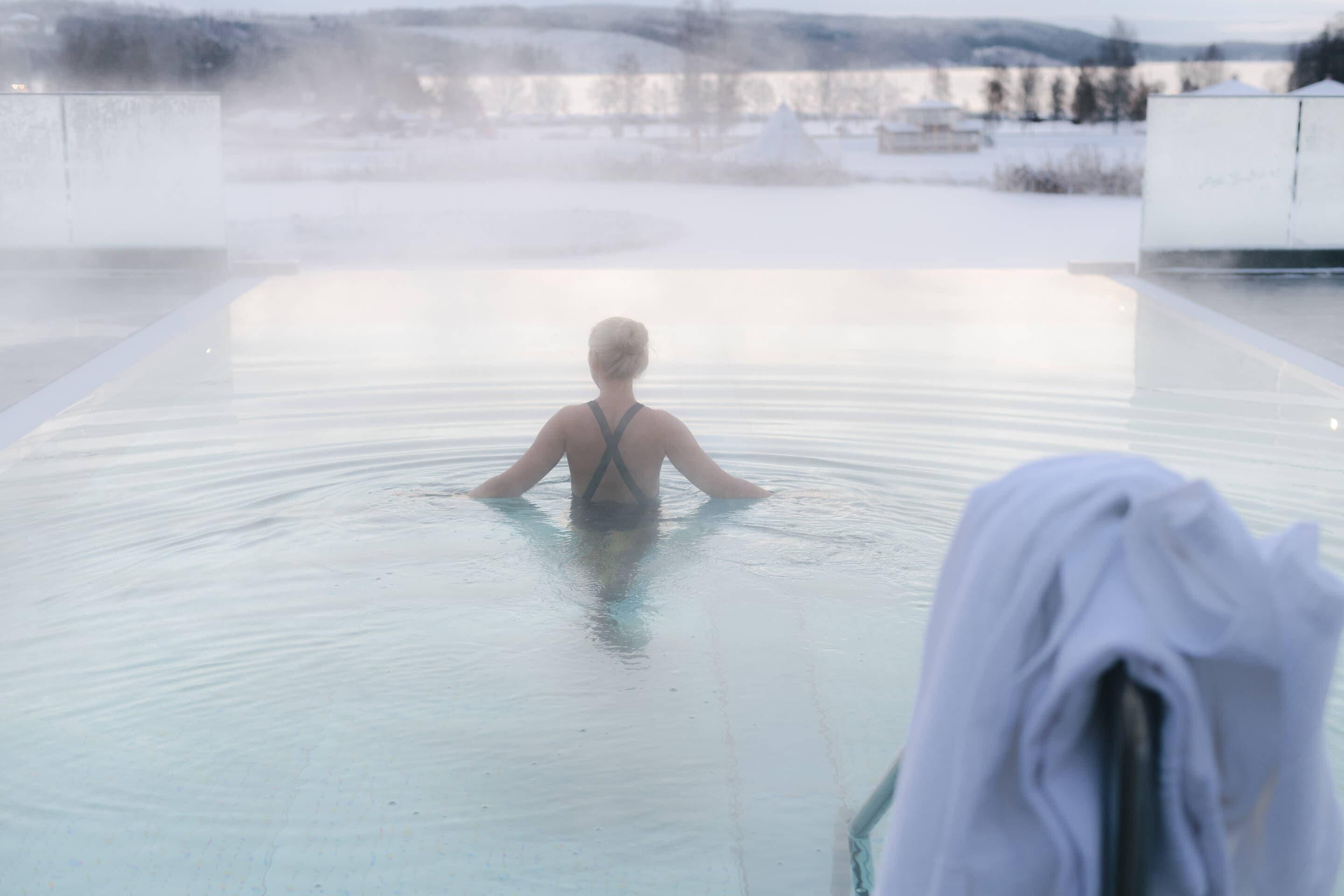 En person i en stor, klar pool med utsikt över ett snötäckt landskap i bakgrunden.