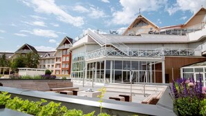 A modern hotel with a glass facade, outdoor seating, and a landscaped garden area. - Selma Spa+ (Sunne)
