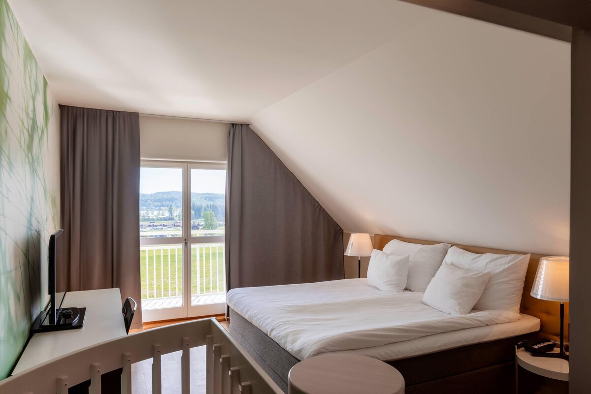 Habitación de hotel con cama, escritorio, silla, lámpara y vistas a un balcón.