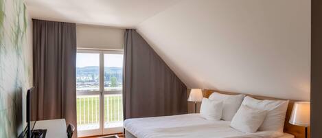 A hotel room with a bed, a desk, a chair, a lamp, and a view of a balcony.