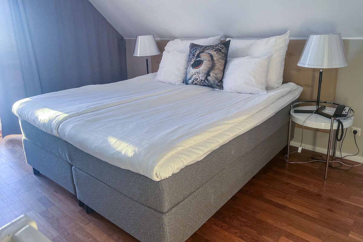 A neatly made bed with white linens and a grey headboard, a bedside lamp, and a power outlet on the nightstand.