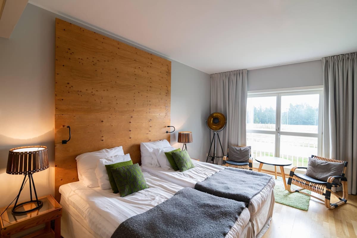 A hotel room with a wooden headboard, a bed with white linens and green pillows, a side table with a lamp, a small round table, a wicker chair, and a view of the outdoors through a large window.