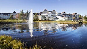 A hotel with a large pond and a fountain in the foreground, surrounded by well-maintained lawns and trees. - Selma Spa+ (Sunne)