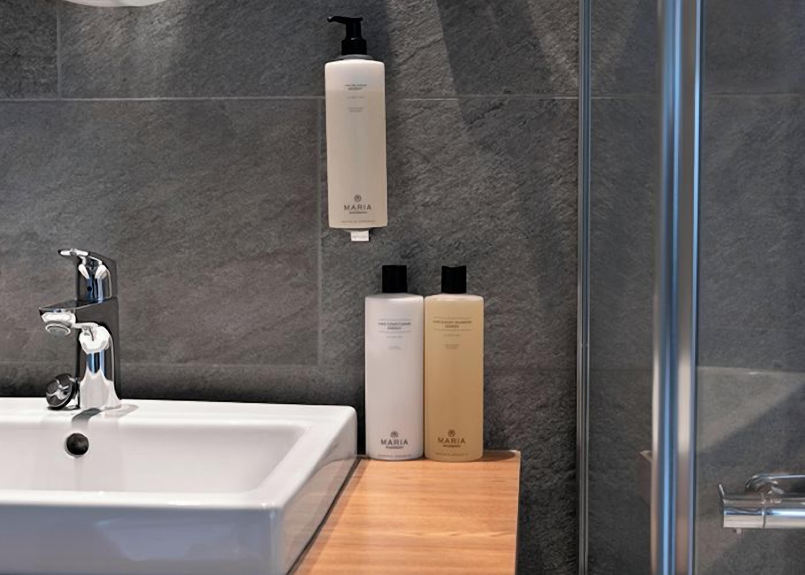 A bathroom with a white sink, a glass shower enclosure, and two bottles of Maria brand products on the countertop.