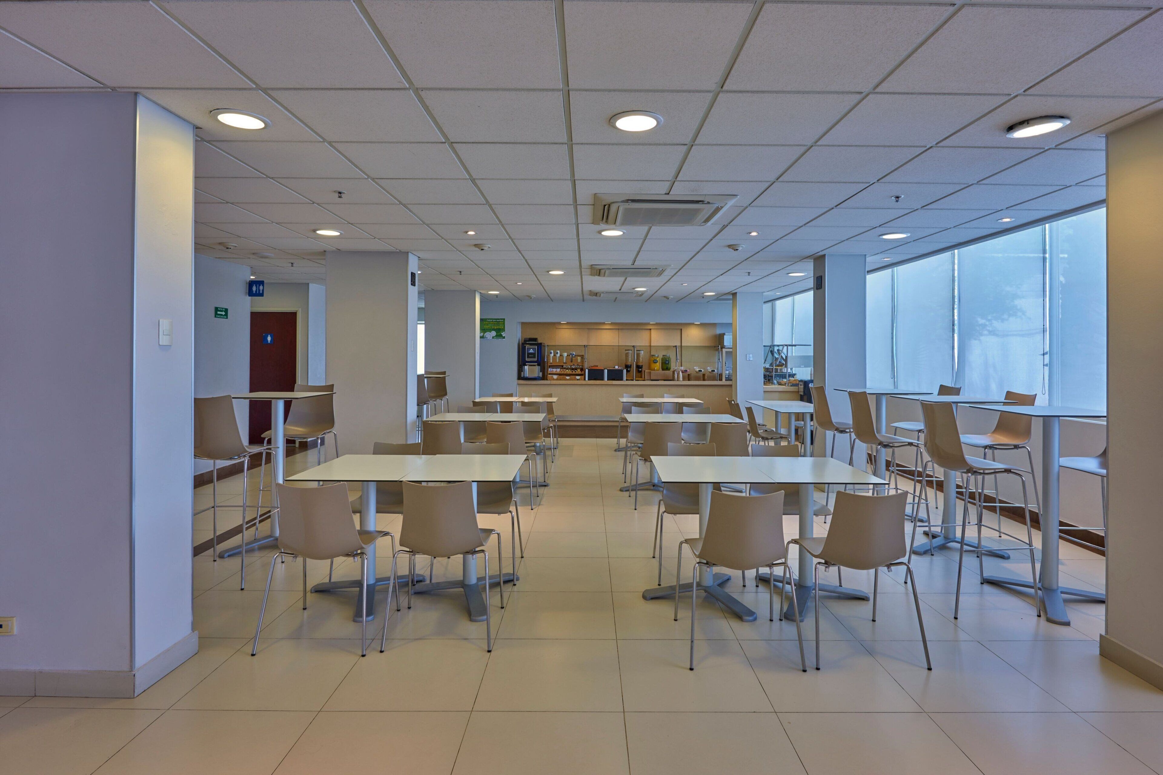 A spacious cafeteria with multiple tables and chairs, large windows, and a kitchen area in the background.
