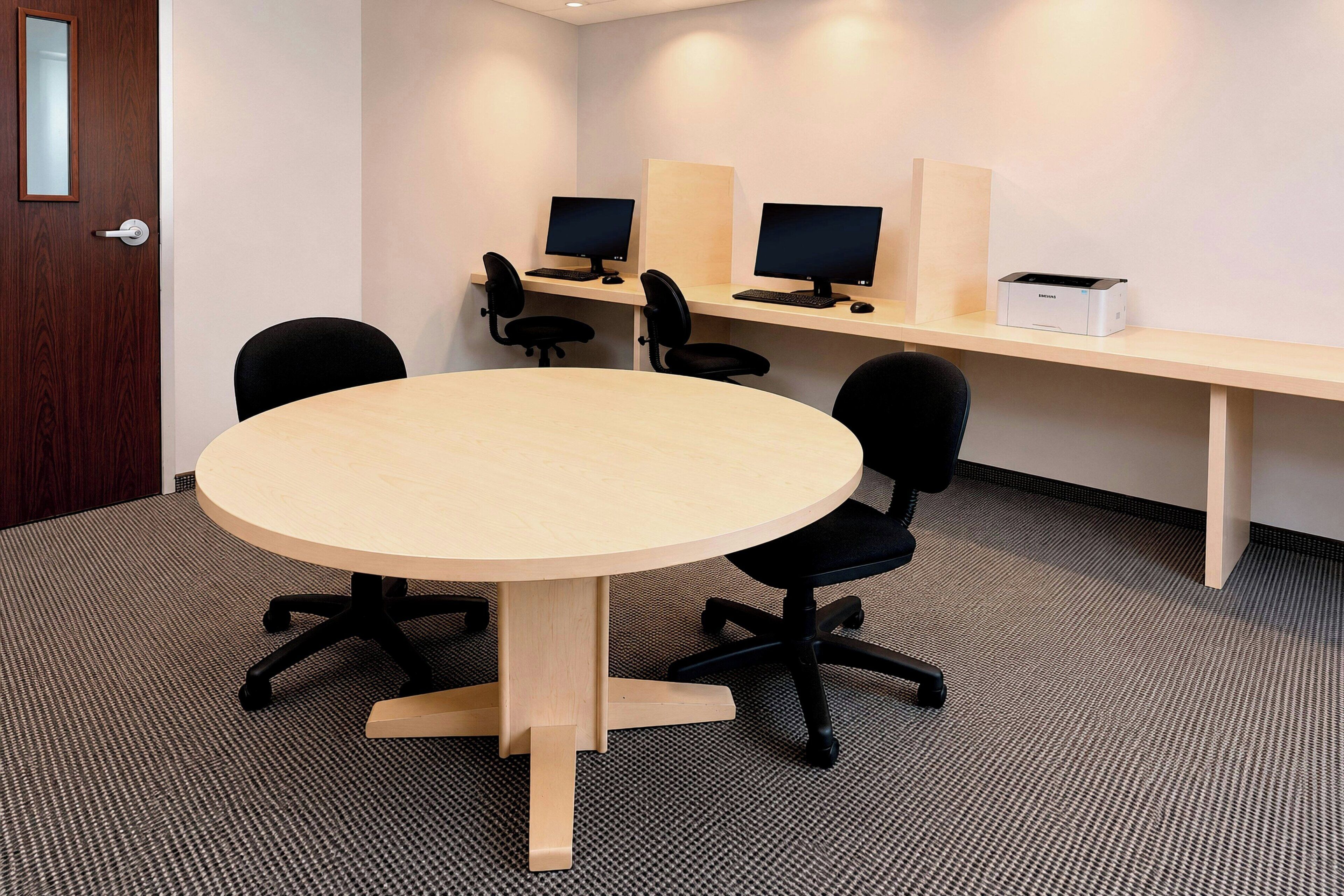 A conference room with a round table, four black office chairs, two computer desks with monitors, and a printer.