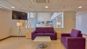 Un vestíbulo moderno con un sofá morado, una mesa con cubierta de vidrio, un televisor de pantalla plana y una recepción.