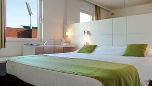 A hotel room with a large bed, a bedside table, a lamp, and a window with a view of a building. - Hotel Expo (Brussels)