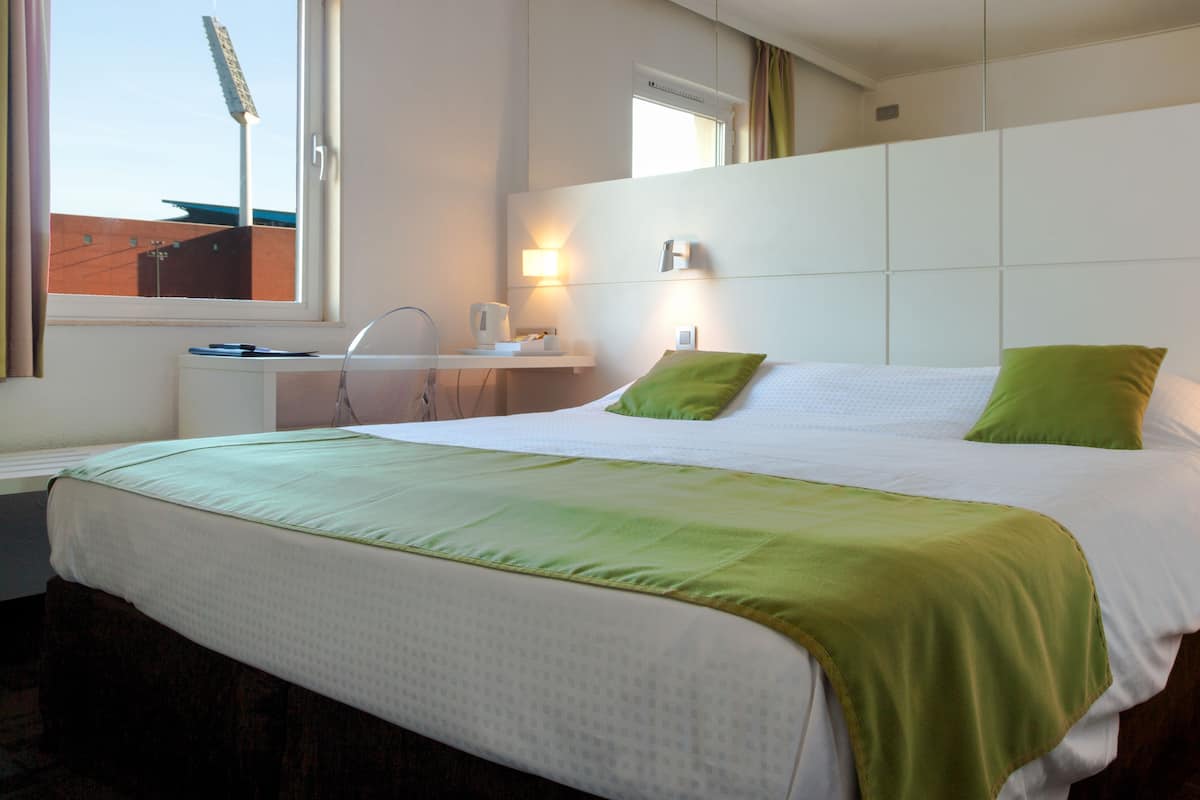 A hotel room with a large bed, a bedside table, a lamp, and a window with a view of a building.