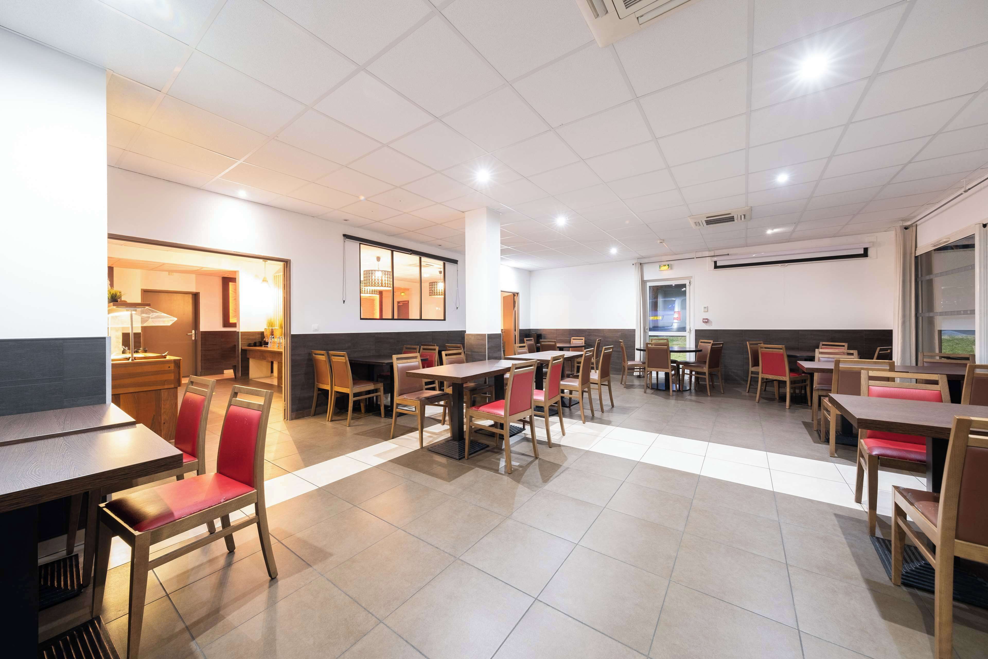 A spacious dining area with multiple tables and chairs, a kitchen area in the background, and a large window providing natural light.