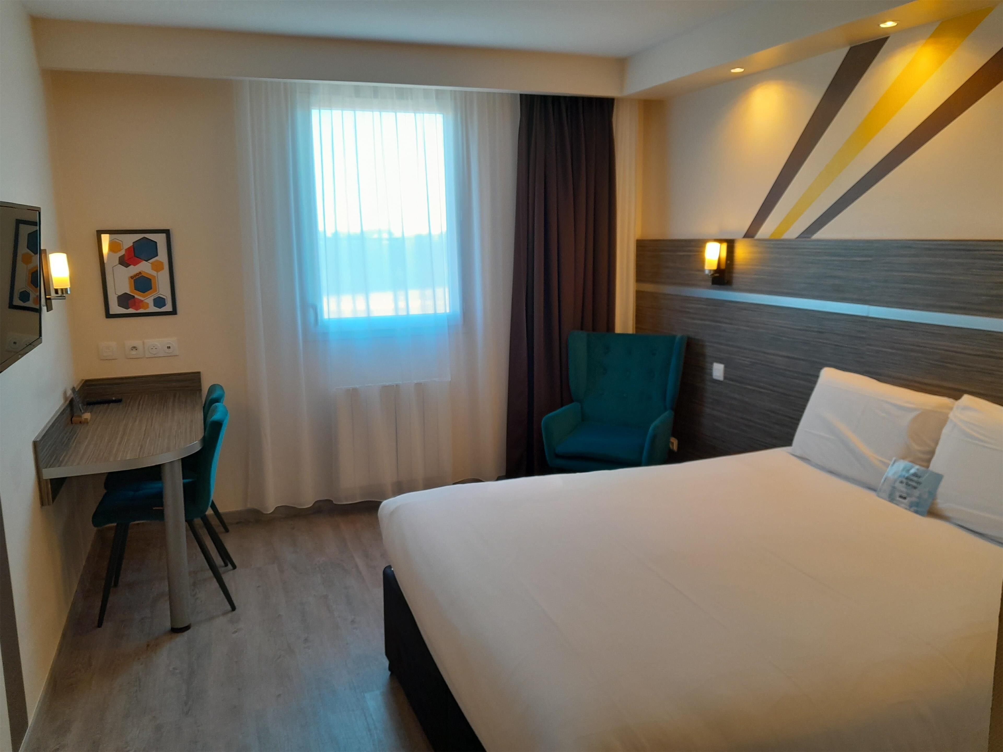 A hotel room with a bed, a desk with a chair, a window with curtains, and a wall-mounted light fixture.