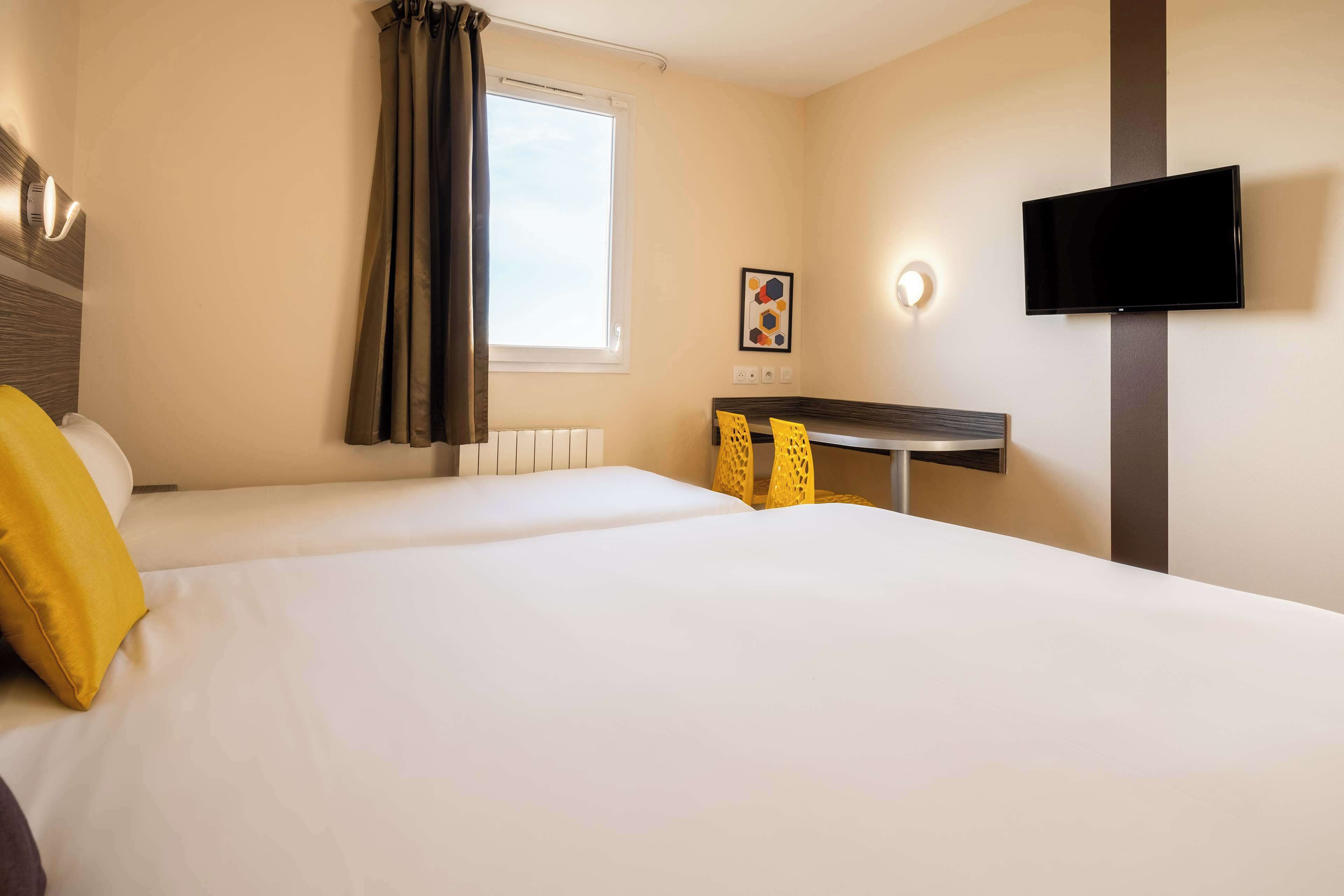 A hotel room with two beds, a desk, a flat-screen TV, and a window with curtains.