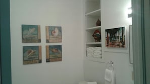 A bathroom with four framed artworks on the wall, a white towel rack, and a shelf with folded towels. - Astor Bridge Marina & Motel (Astor)