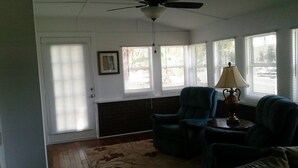 A room with a ceiling fan, a window with blinds, a chair, a table with a lamp, and a rug. - Astor Bridge Marina & Motel (Astor)