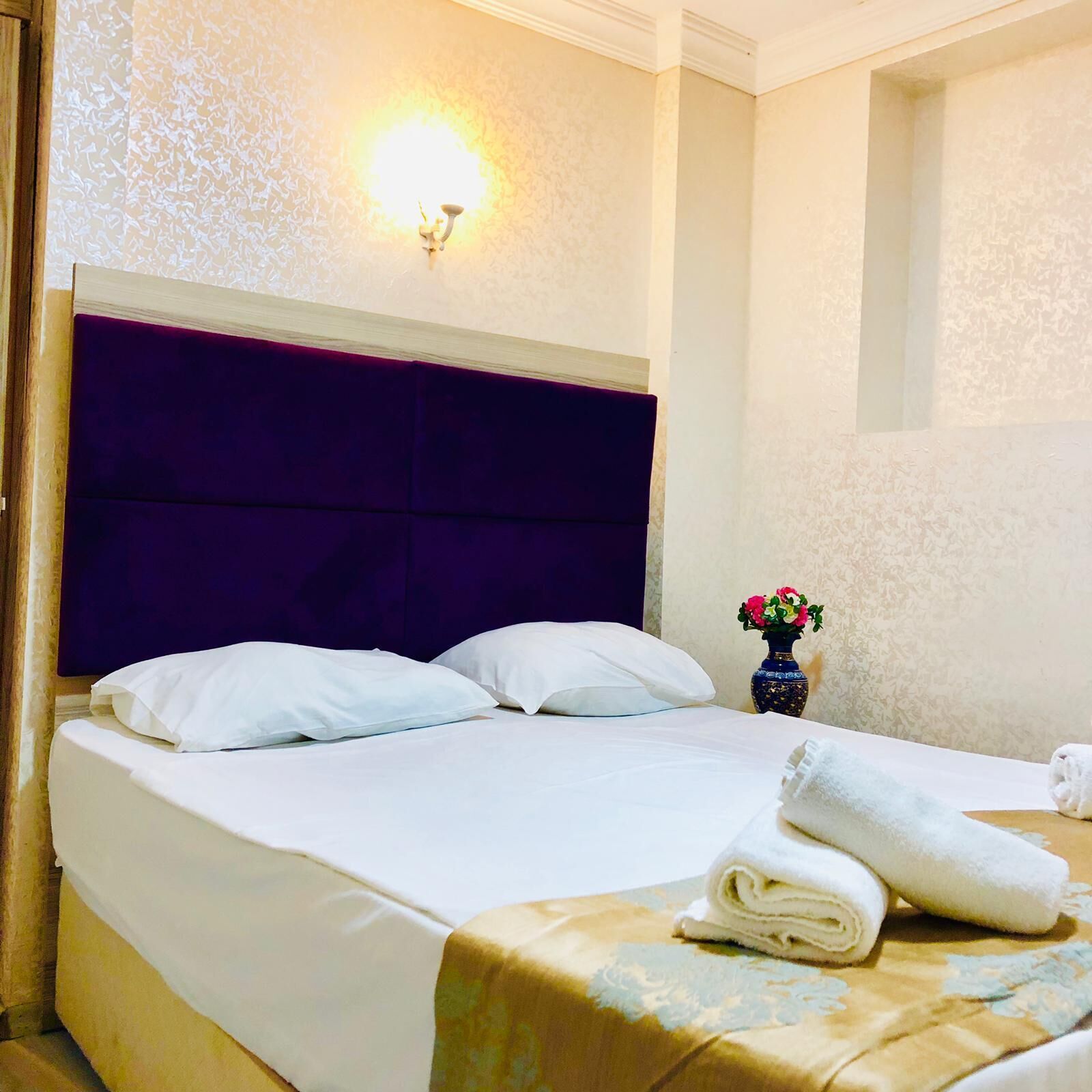 A hotel room with a large bed, white bedding, a purple headboard, a wall-mounted light, and a vase with flowers.