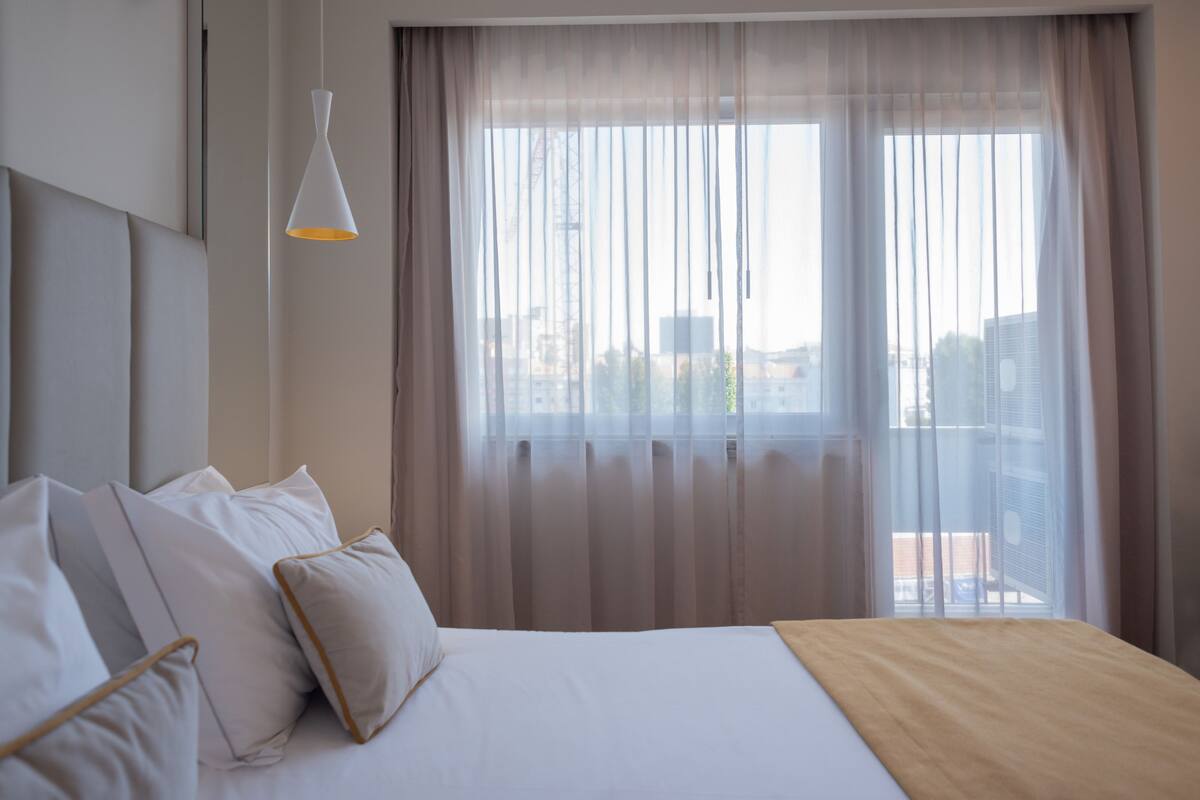 Et hotelværelse med en seng, hvide sengetøj, en hængende lampe, lette gardiner og udsigt til bygninger gennem vinduet.