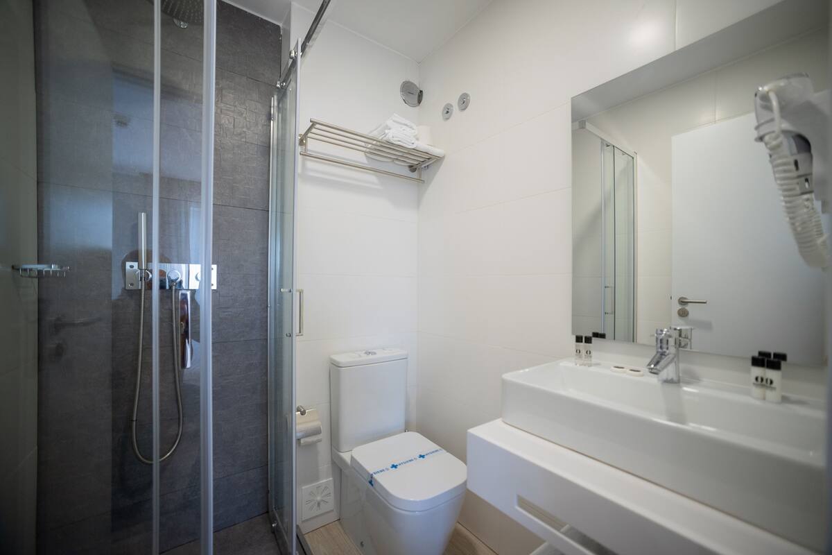 Et moderne badeværelse med en bruseniche med glasvægge, et hvidt toilet og et stort spejl over en hvid vask.