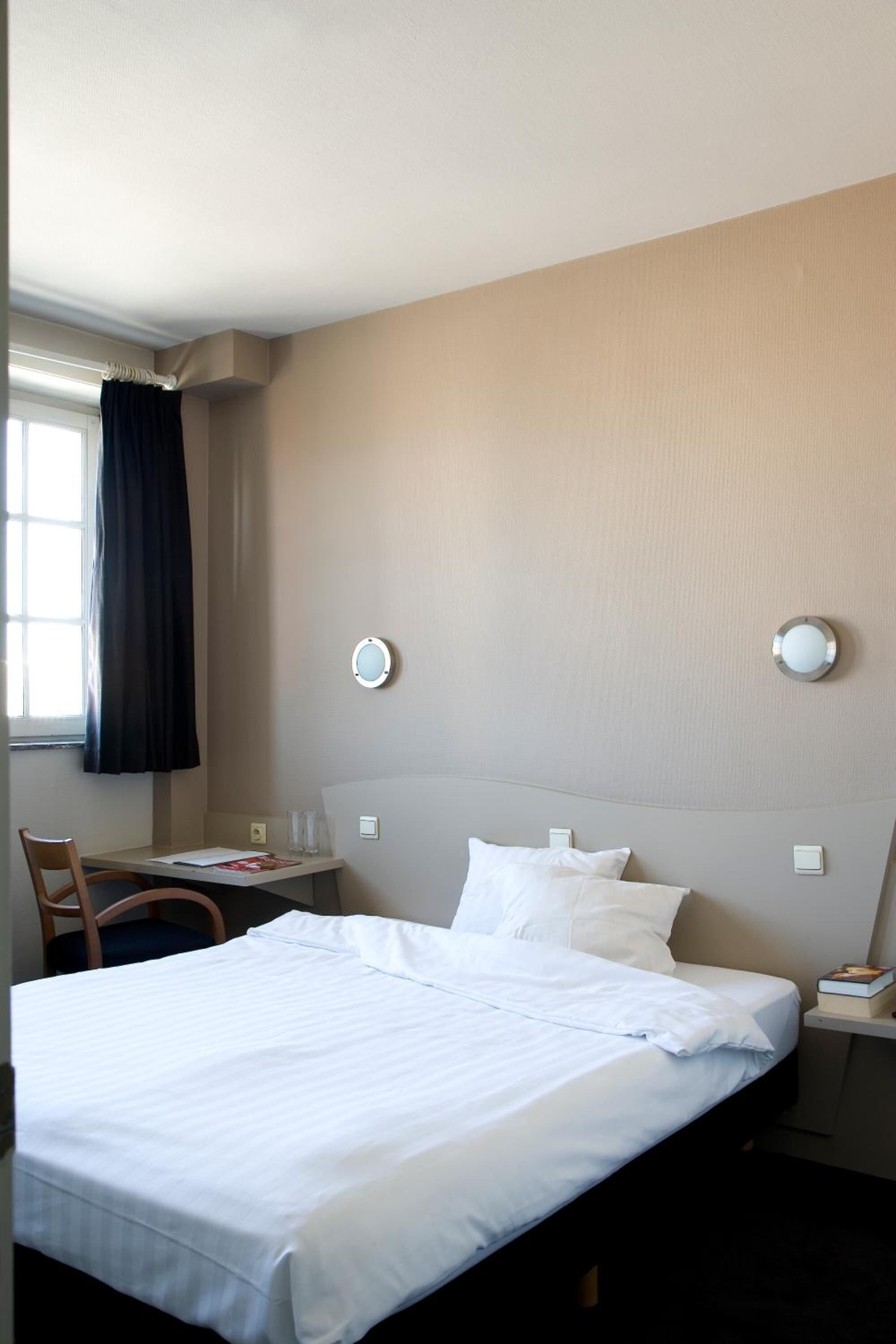 A hotel room with a bed, a desk, a chair, and a window with curtains.