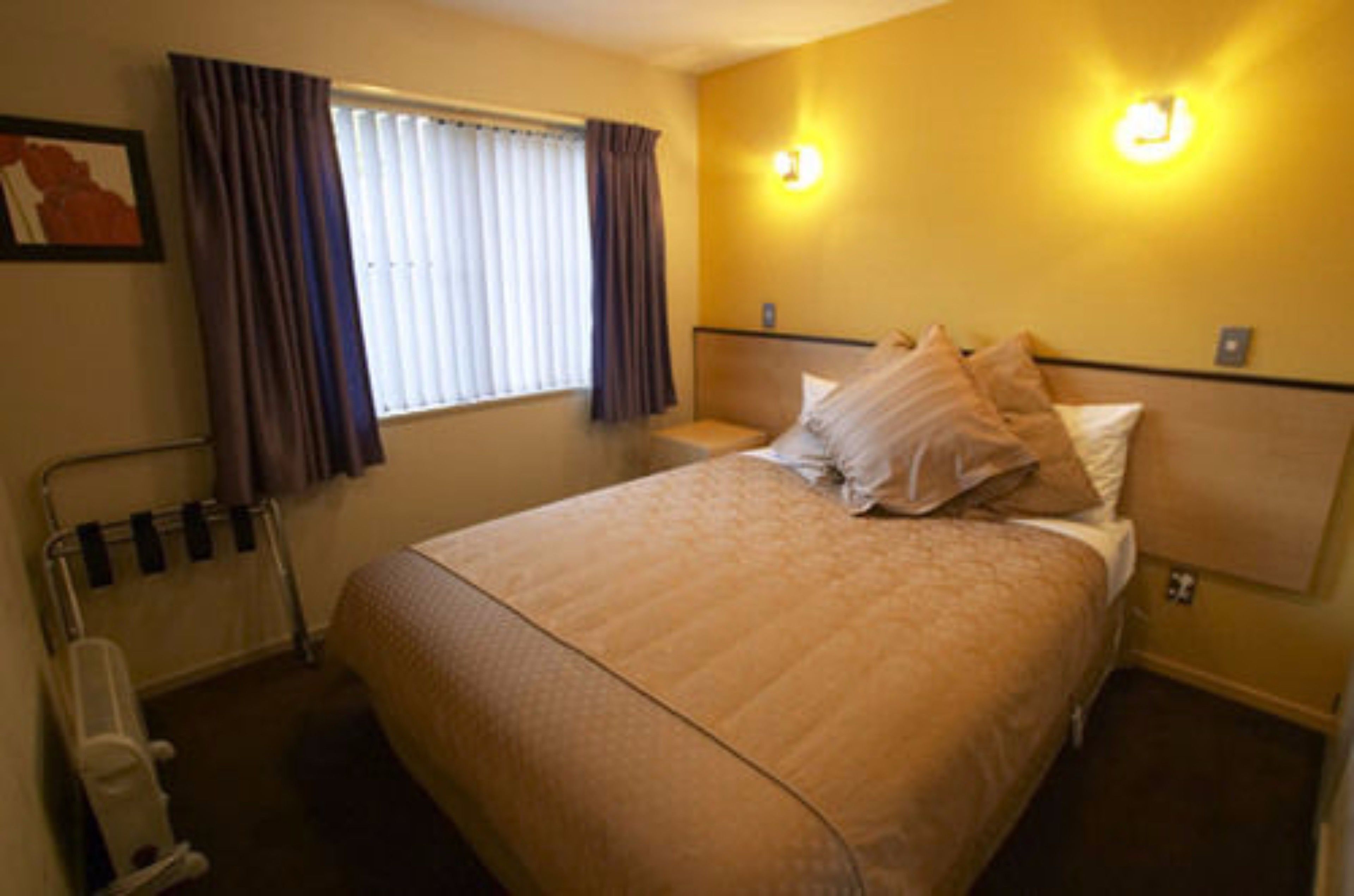 A hotel room with a bed, a bedside table, a lamp, a window with curtains, and a wall-mounted light fixture.