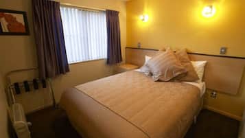 A hotel room with a bed, a bedside table, a lamp, a window with curtains, and a wall-mounted light fixture.