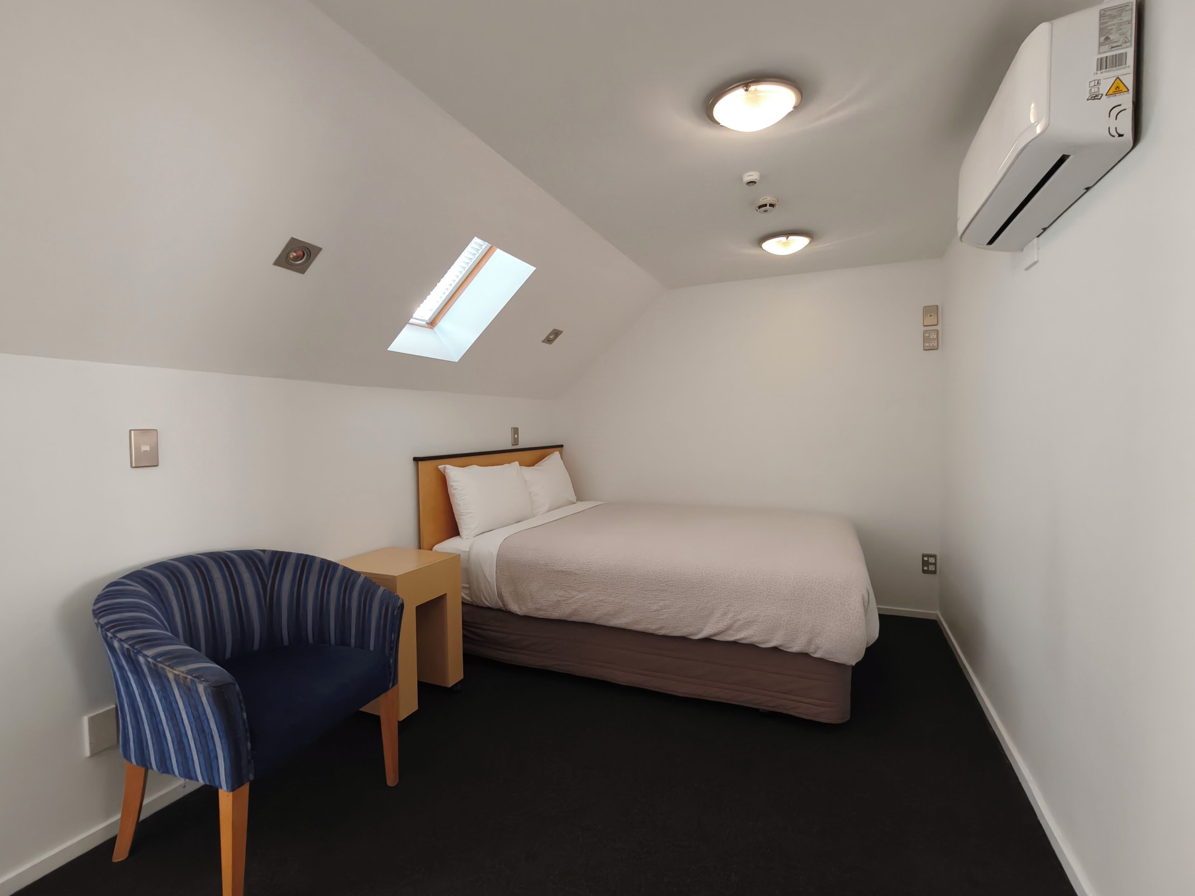 Habitación de hotel pequeña y de una sola habitación con una cama, una silla, una mesita, un tragaluz y un equipo de aire acondicionado.
