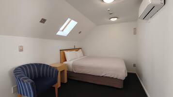 Habitación de hotel pequeña y de una sola habitación con una cama, una silla, una mesita, un tragaluz y un equipo de aire acondicionado.
