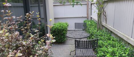 Een terras met een ronde tafel en twee stoelen, omgeven door groen en een houten hek.