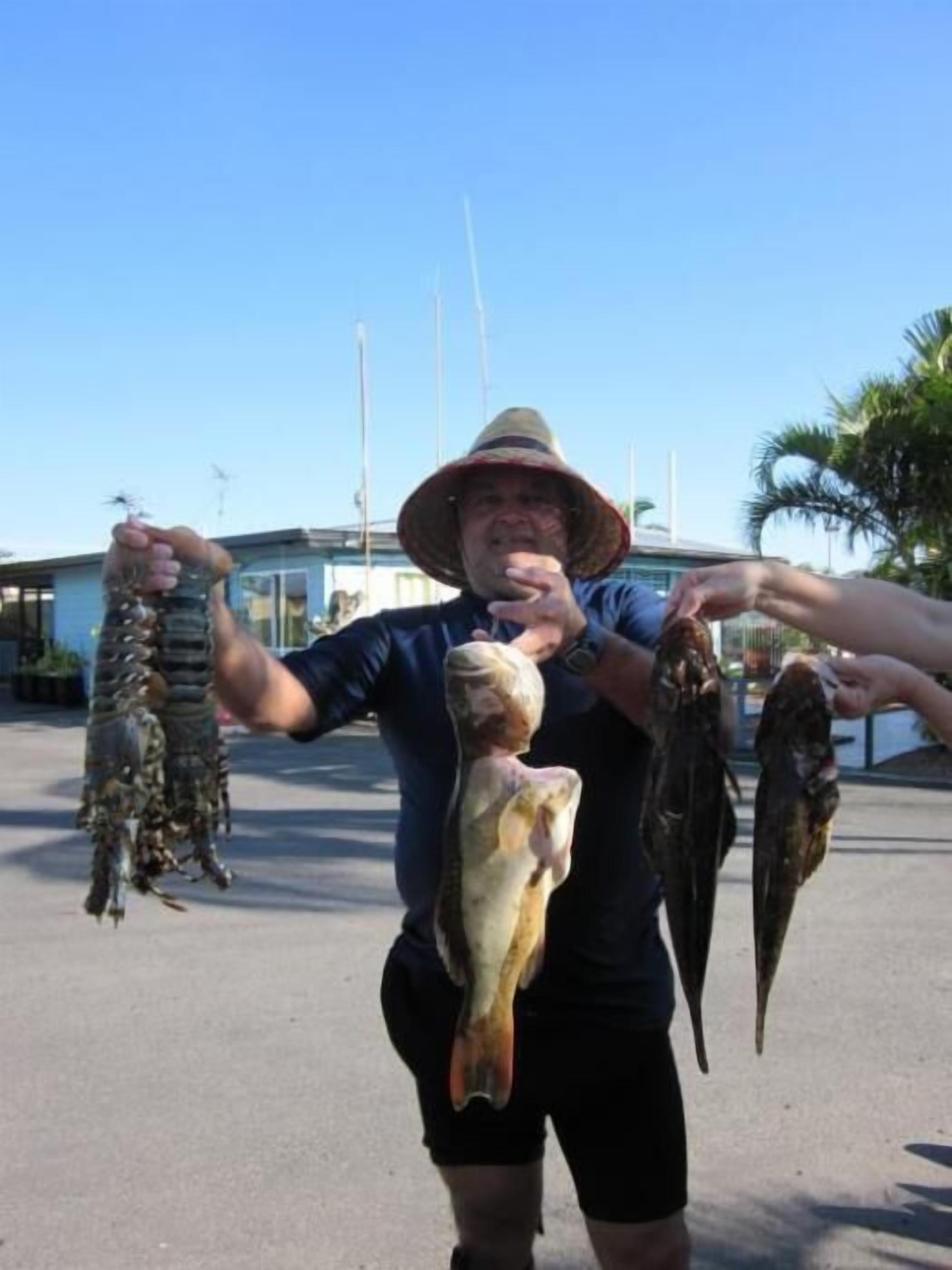 A person holding a lobster and a fish, with a building and palm trees in the background.