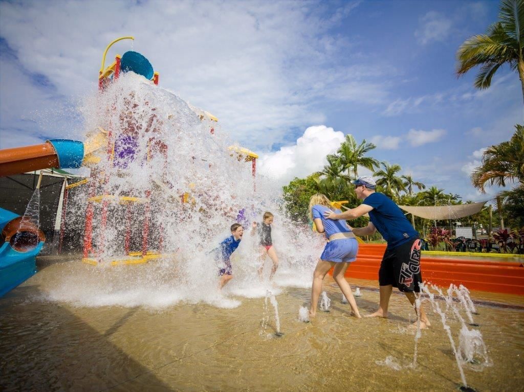 A water play area with a large water fountain and slides, surrounded by palm trees and a clear sky.
