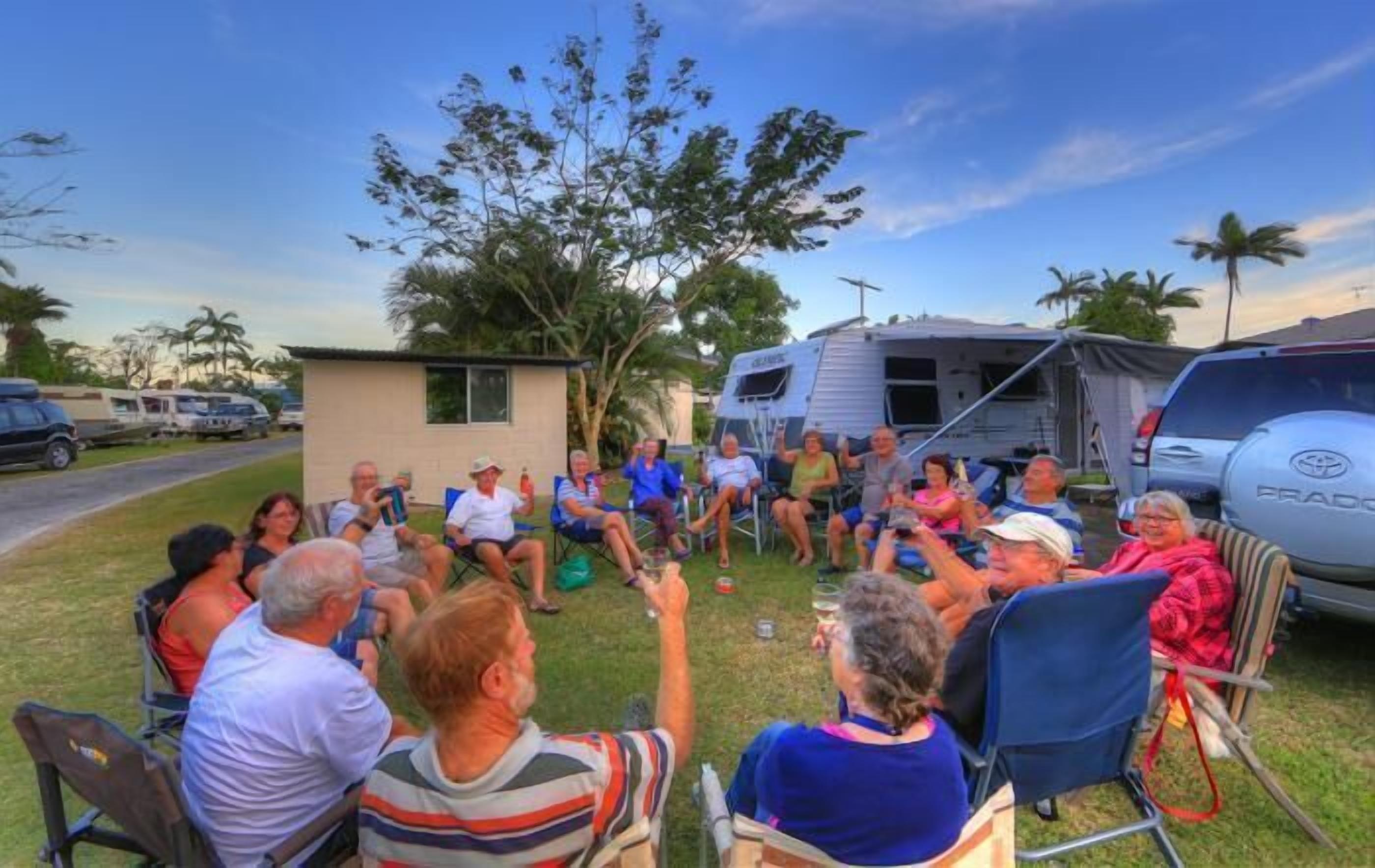 A group of people seated outdoors in lawn chairs, with a trailer and parked cars in the background.