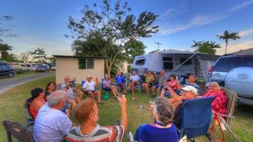 A group of people seated outdoors in lawn chairs, with a trailer and parked cars in the background.