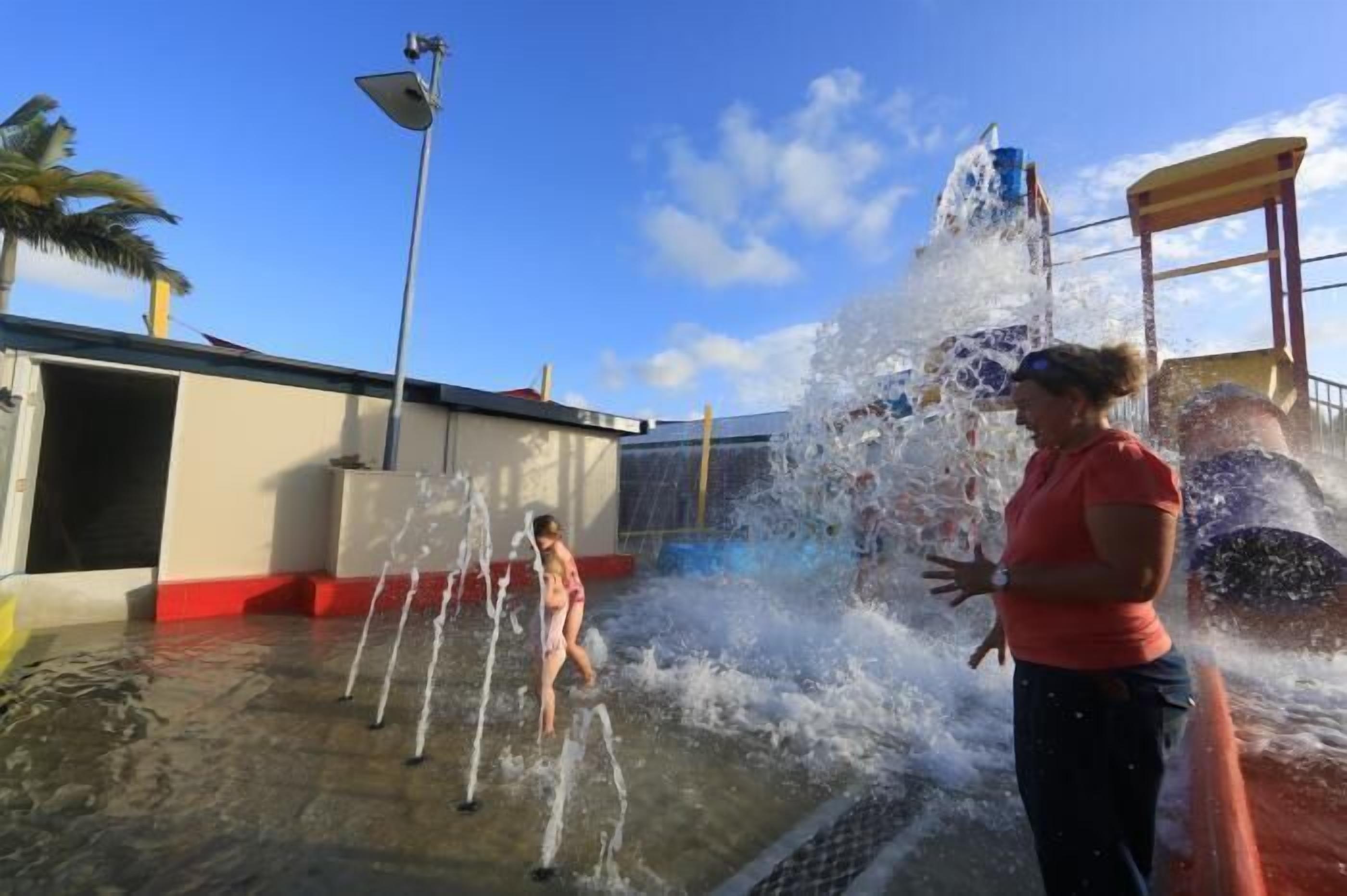 A water play area with multiple water jets and a slide, surrounded by a building with a red and white facade.