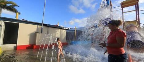 A water play area with multiple water jets and a slide, surrounded by a building with a red and white facade.