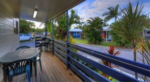 A covered outdoor area with a wooden deck, a table and chairs, and a view of a parking lot and buildings in the background. - Kurrimine Beach Holiday Park (Kurrimine Beach)