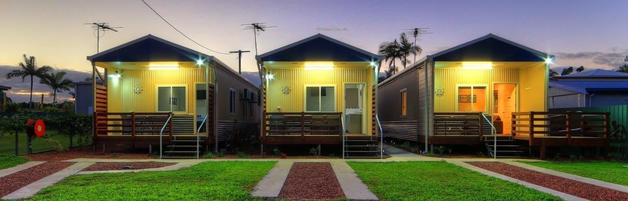 Three small, single-story cabins with a porch and a small staircase, each with a door and windows, set in a grassy area with a clear sky.