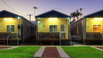 Three small, single-story cabins with a porch and a small staircase, each with a door and windows, set in a grassy area with a clear sky.