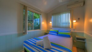 Un dormitorio con cama, ropa de cama a rayas, una ventana con persianas y una mesita de noche.