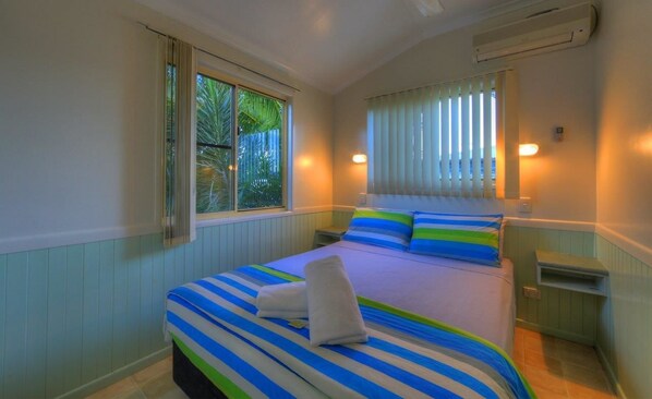 A bedroom with a bed, striped bedding, a window with blinds, and a nightstand.